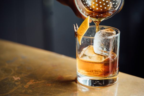 A cocktail being strained over a large ice cube in a rocks glass, garnished with an orange peel, on a bar top.