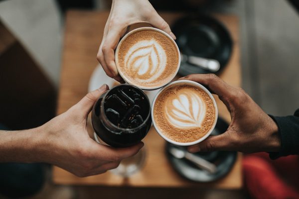 Overhead view of two hands holding latte-art coffees and a third holding an iced black drink over a wooden table.