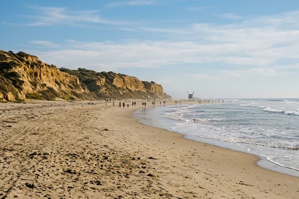 A long sandy beach below golden sandstone bluffs on the Southern California coast, with a lifeguard tower in the distance and small figures walking near gentle surf.