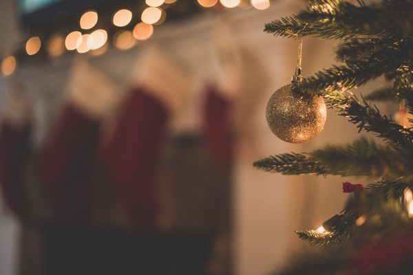 A gold glitter ornament hanging on a Christmas tree in front of softly blurred string lights and stockings.